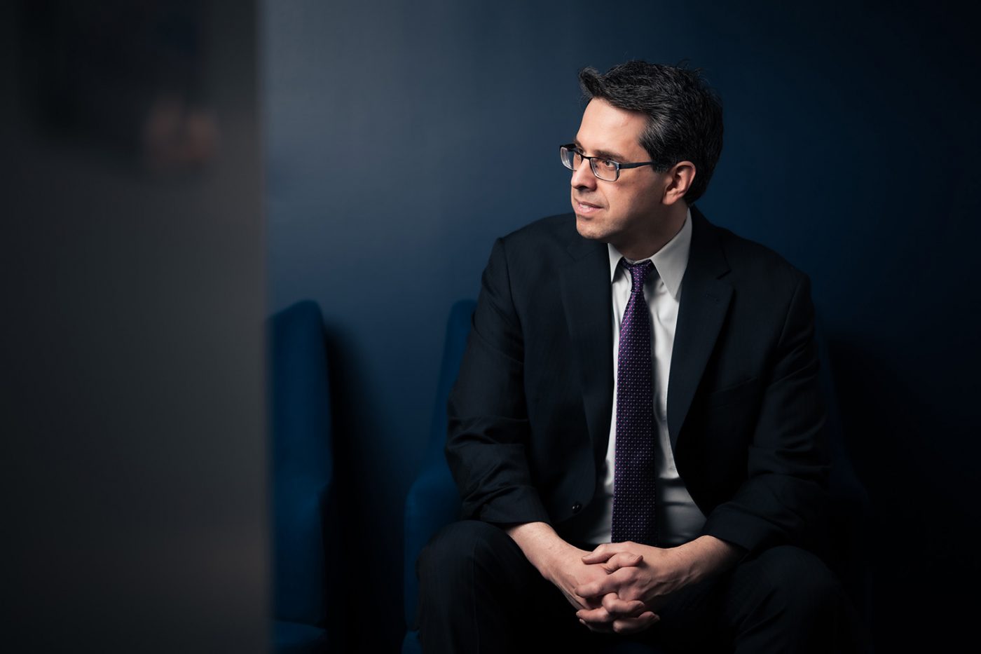 Picture of Robert Bird, wearing a black suit, sitting and looking slightly to the side, with hands folded together in lap. Background color is dark blue.